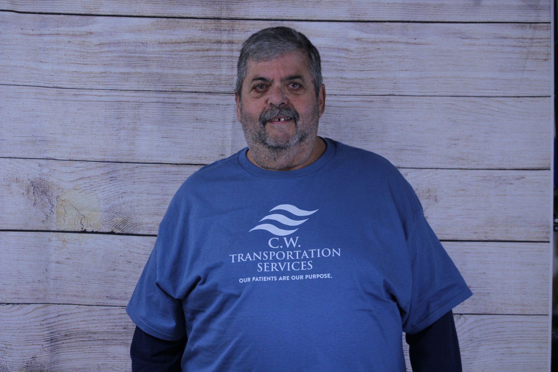 A person with short hair and a beard wearing a blue C.W. Transportation Services t-shirt against a wood-paneled background.