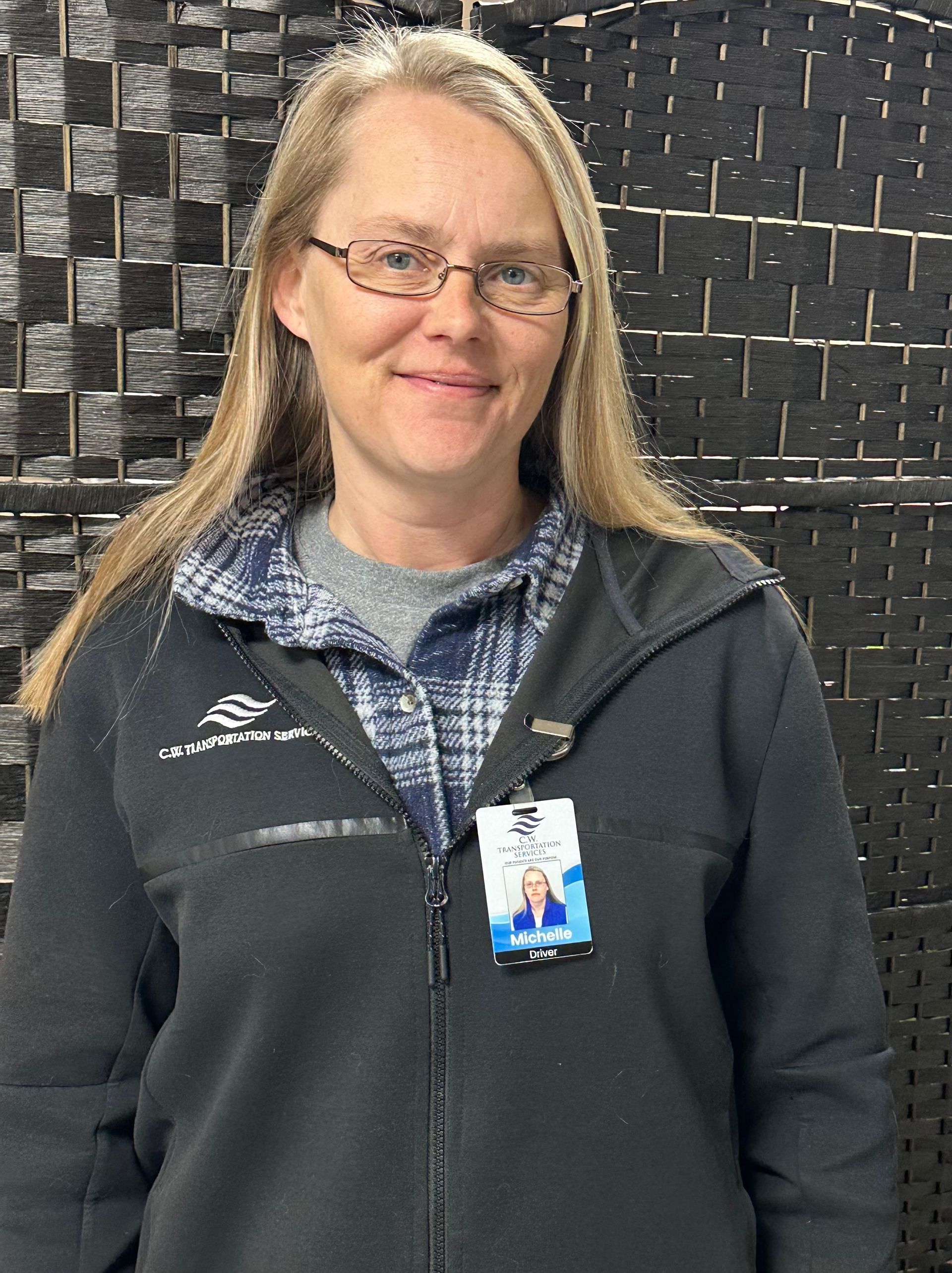 A person with long blonde hair and glasses wearing a black zip-up fleece over a plaid shirt, with a name badge attached.