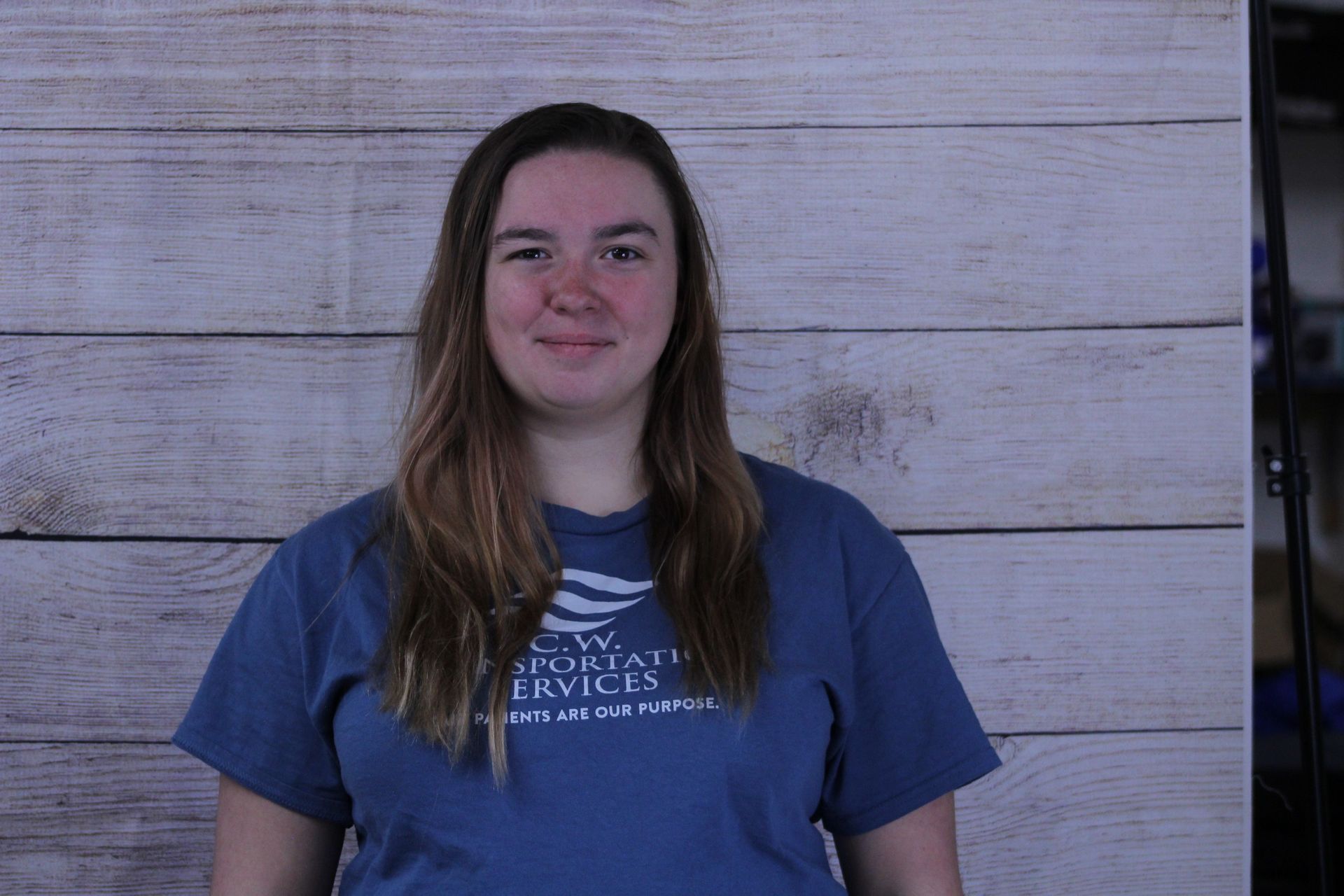 A person with long brown hair, wearing a blue t-shirt, standing in front of a light wood-plank wall.
