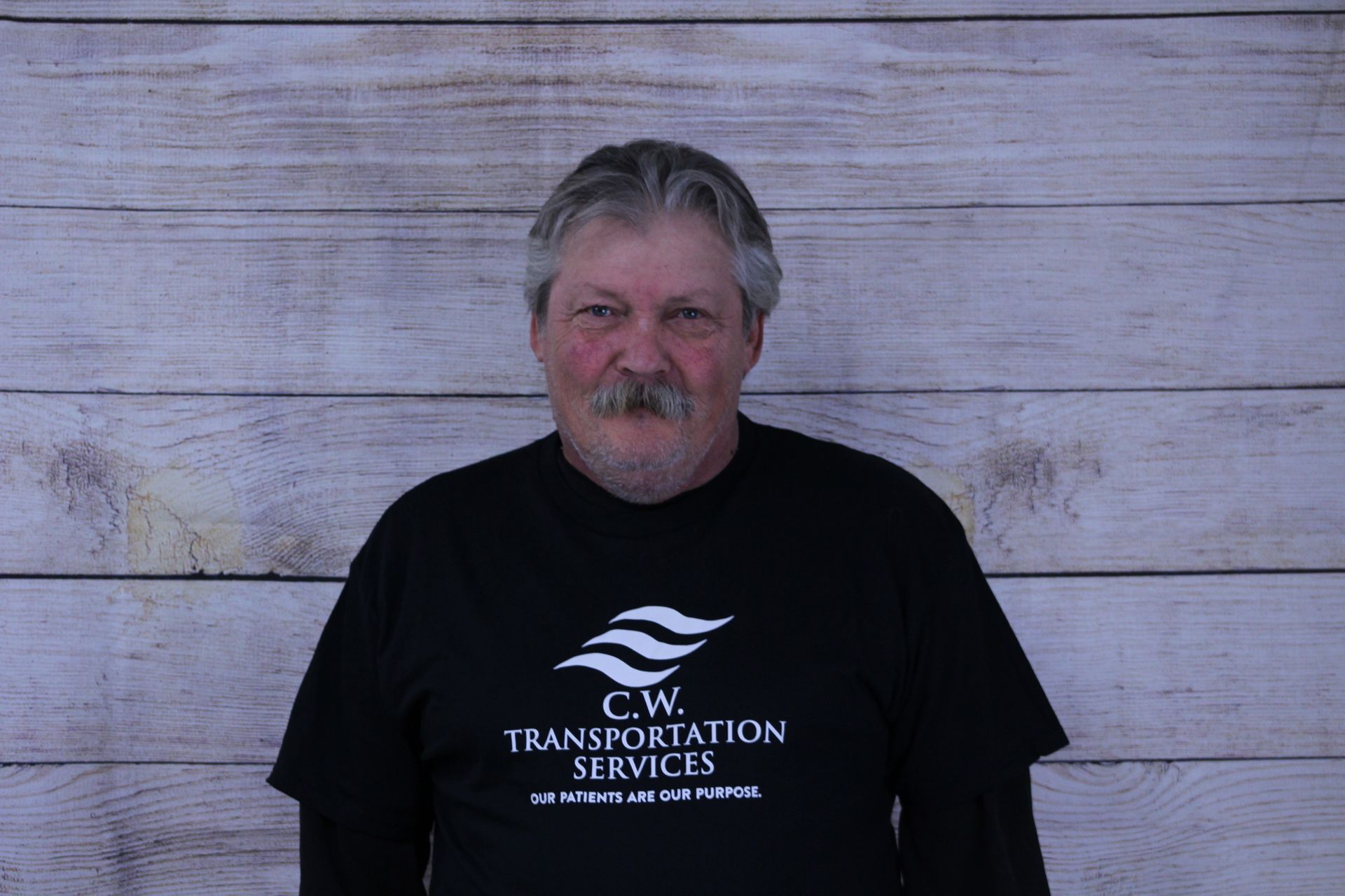 A person with a mustache wearing a black C.W. Transportation Services t-shirt against a light wood-paneled background.