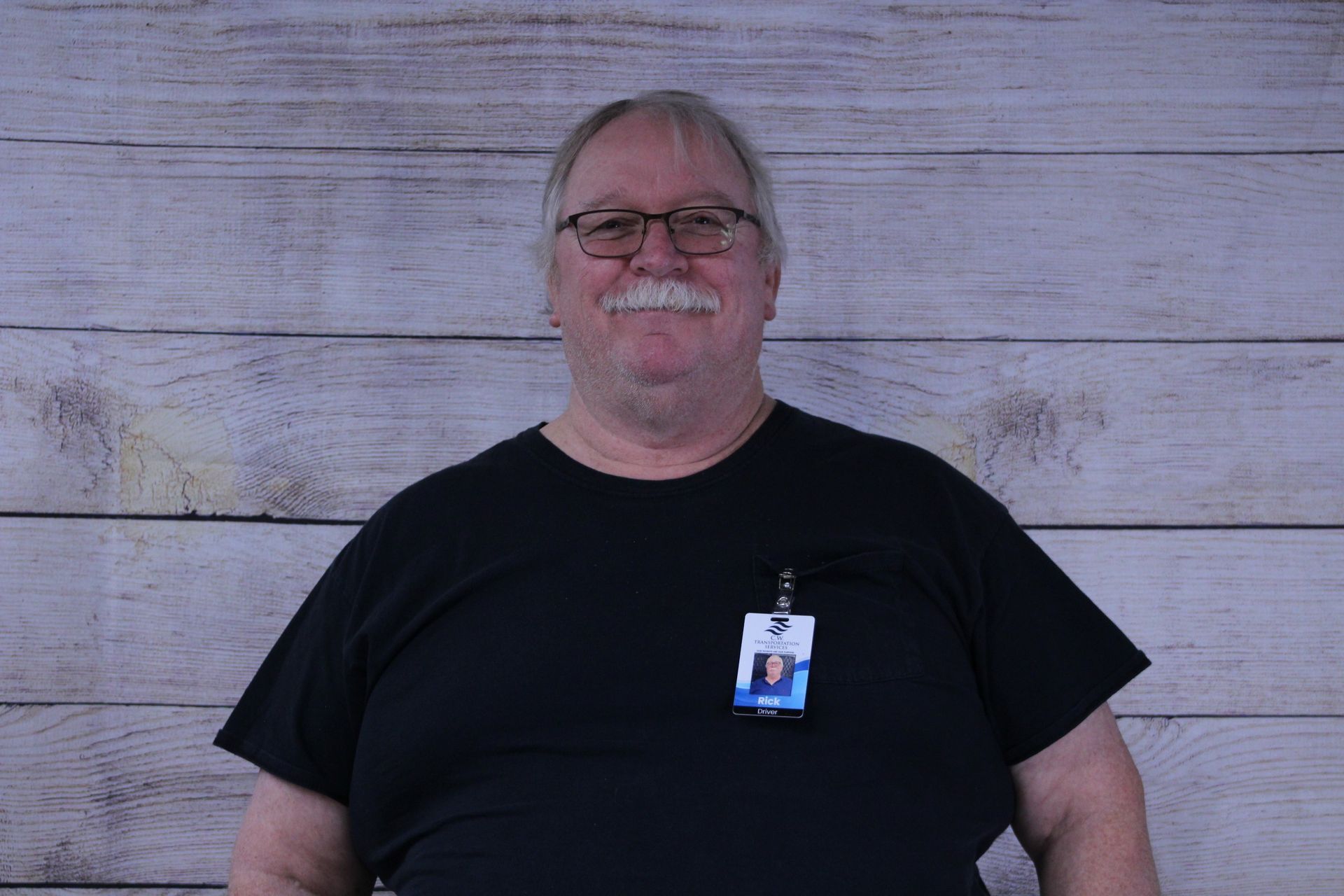 A person with glasses and a mustache wearing a black t-shirt with an ID badge, standing in front of a wood-plank wall.