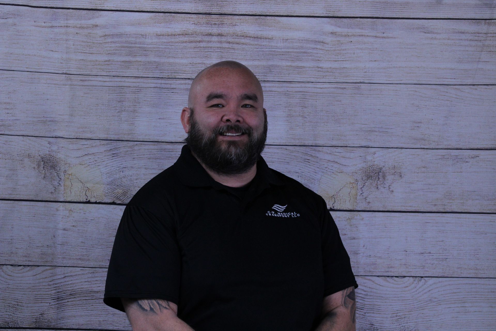 A smiling individual with a beard wearing a black polo shirt stands against a rustic, light-colored wood panel backdrop.
