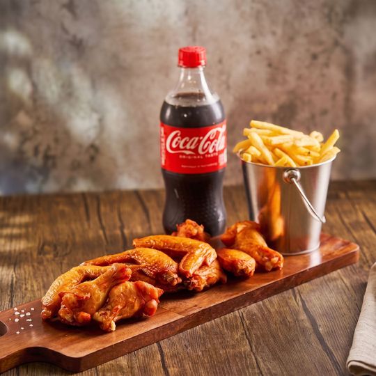 Coca-Cola bottle with fried chicken wings and fries in a metal cup on a wooden board