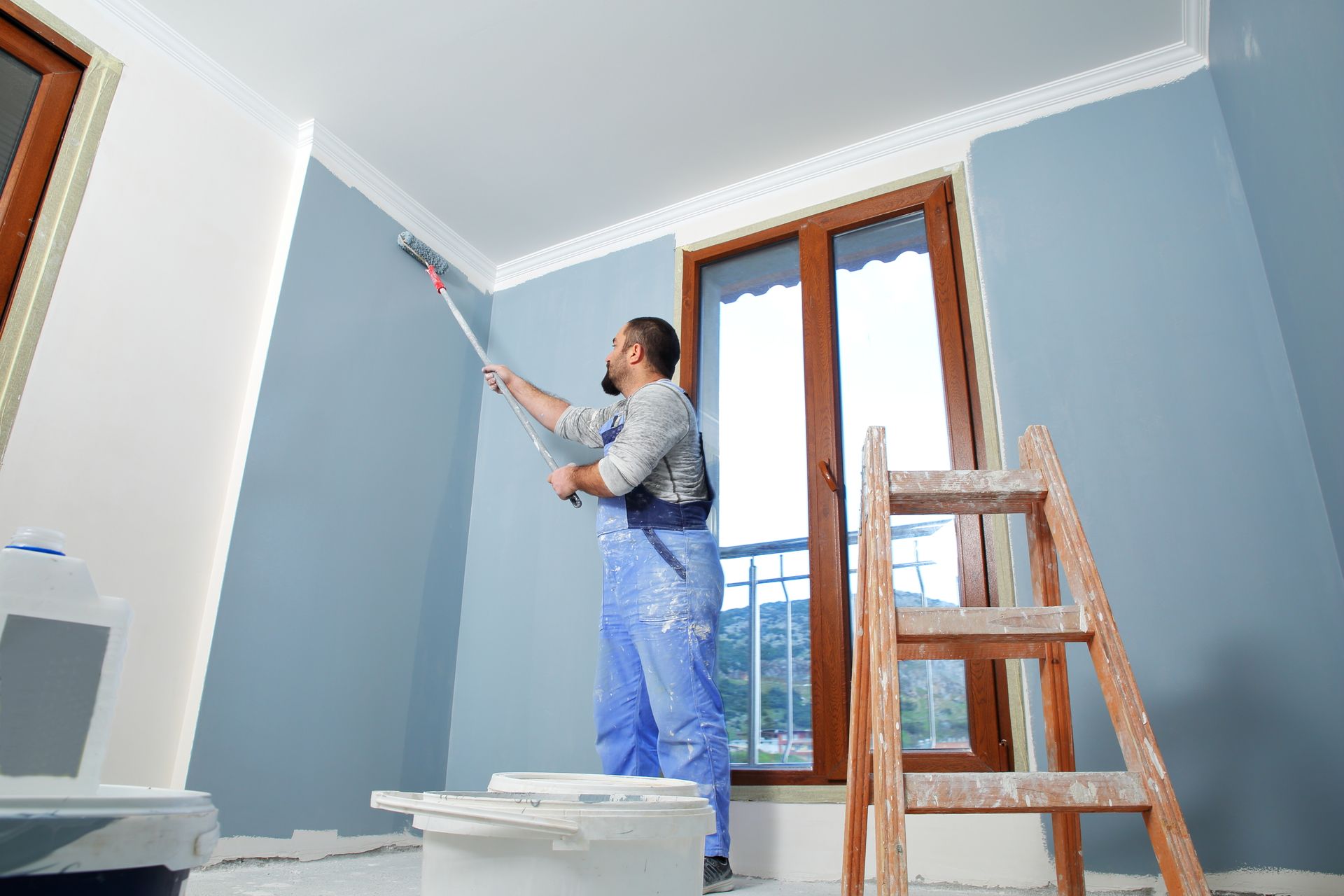 Man in blue coveralls painting a wall blue with a roller, in a room with a wooden window and ladder.