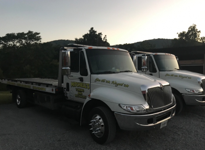 Two white tow trucks parked side by side at dusk, one in front with a flatbed.