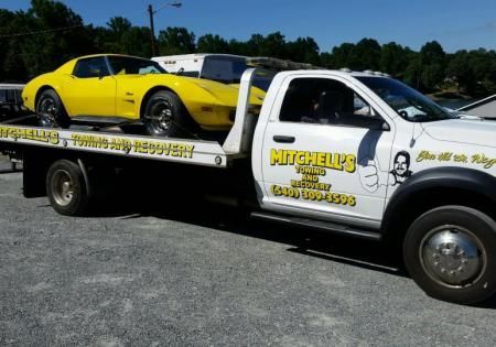 Tow truck carrying a yellow sports car, parked outdoors with Mitchell’s towing logos.