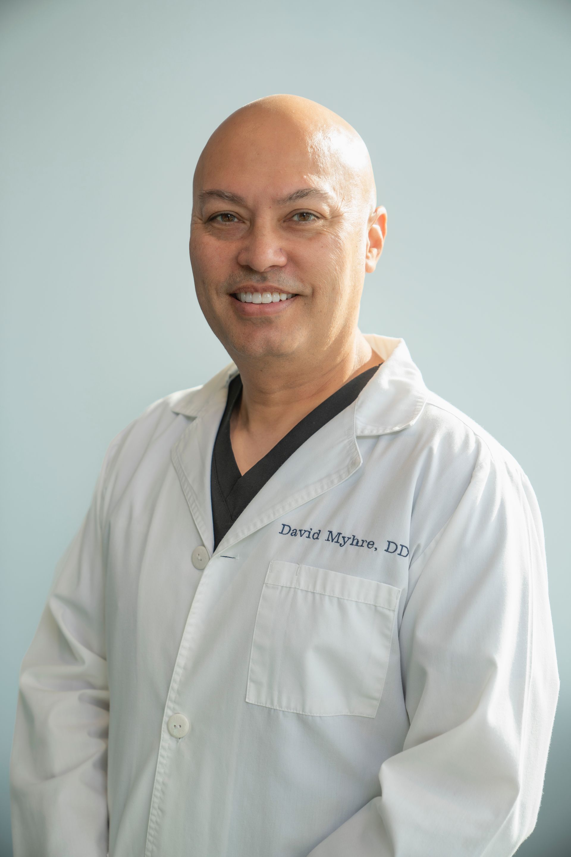 Smiling man in a white lab coat, text on the coat, standing against a light blue backdrop.