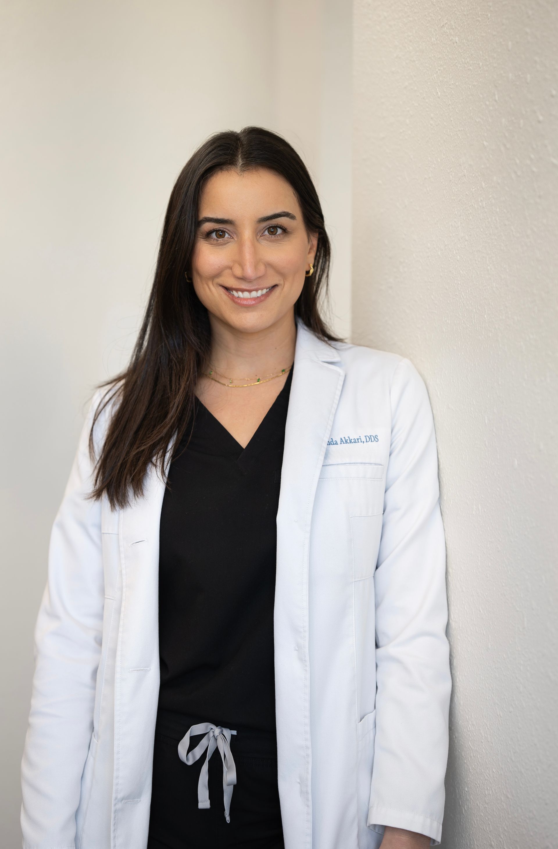 A person wearing a white medical lab coat and black scrubs, smiling while leaning against a light-colored wall.