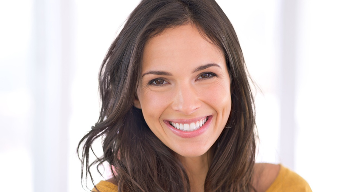 Woman with brown hair smiles, showing white teeth, against a blurred background.