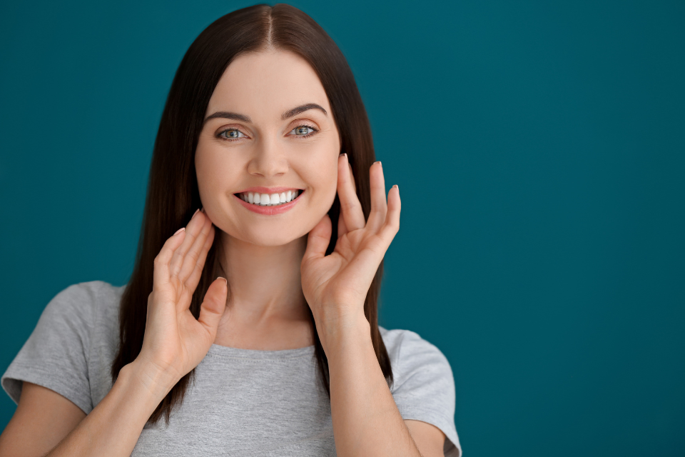 Woman in gray shirt smiles, touching cheeks; teal background.