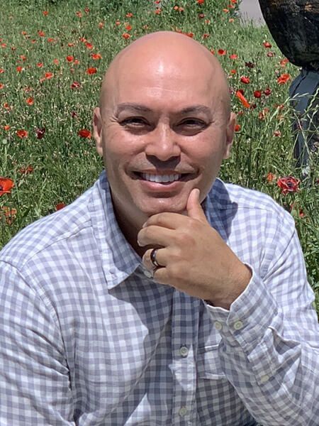 A bald man in a plaid shirt is smiling in front of a field of flowers.