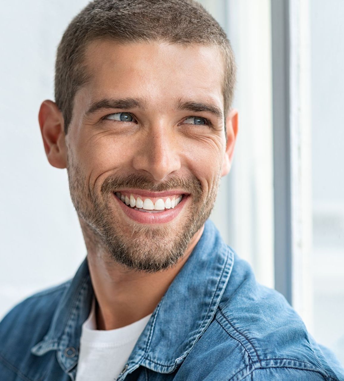 Man with short brown hair smiles, looking to the side; wears denim shirt.