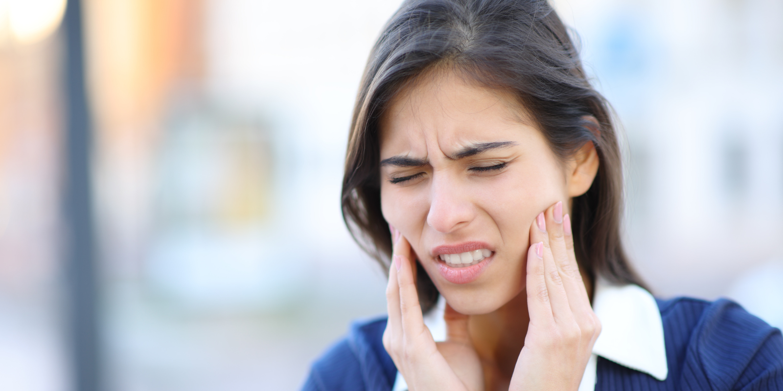 Woman grimacing from TMJ and holding her cheeks, suggesting toothache or facial pain.