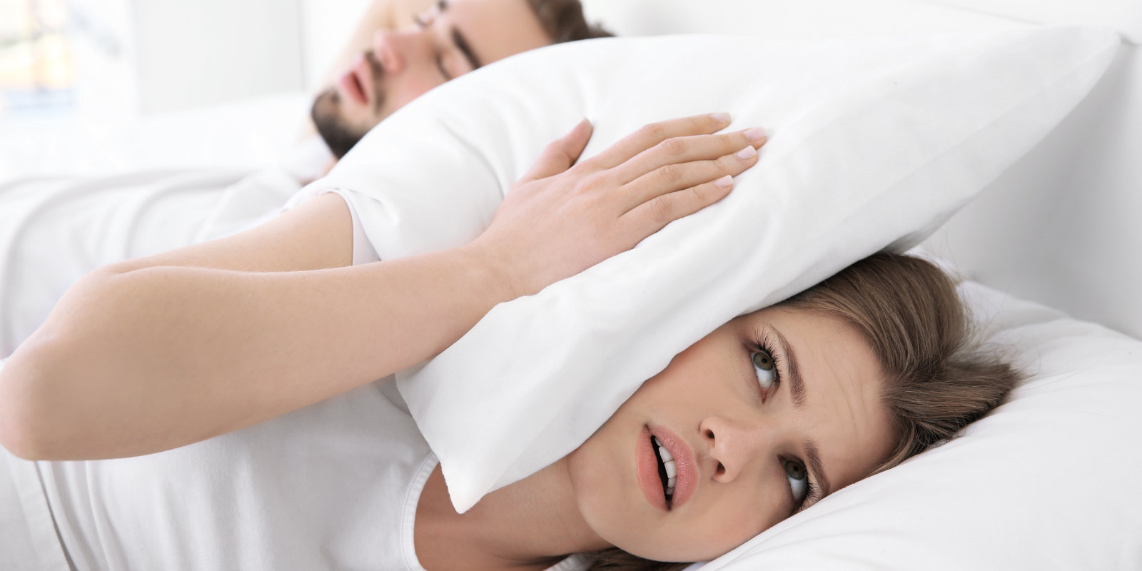 Person lying in bed, holding a pillow over their head and looking stressed or tired