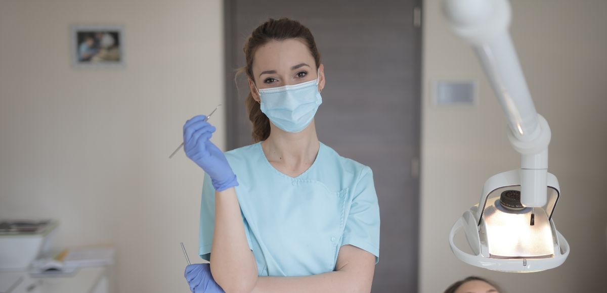 Dental professional in blue scrubs and mask holding a glove beside a dental chair and exam light