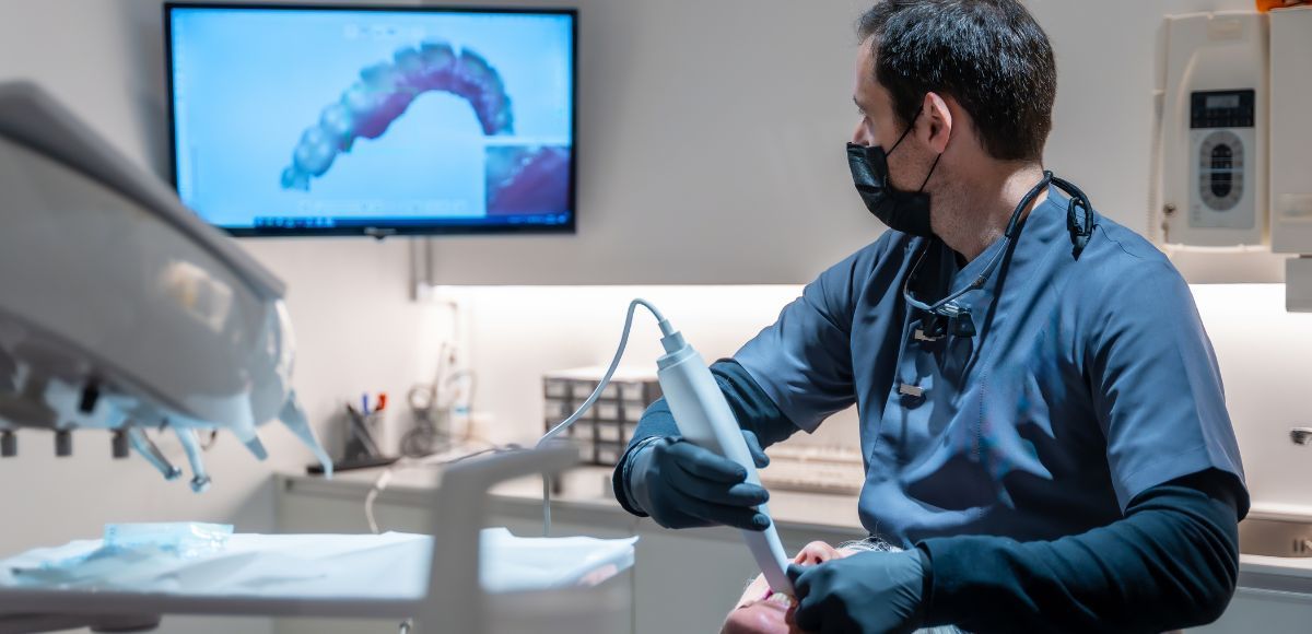 Dentist in blue scrubs seated at a dental chair, with a monitor showing a tooth scan in the background.