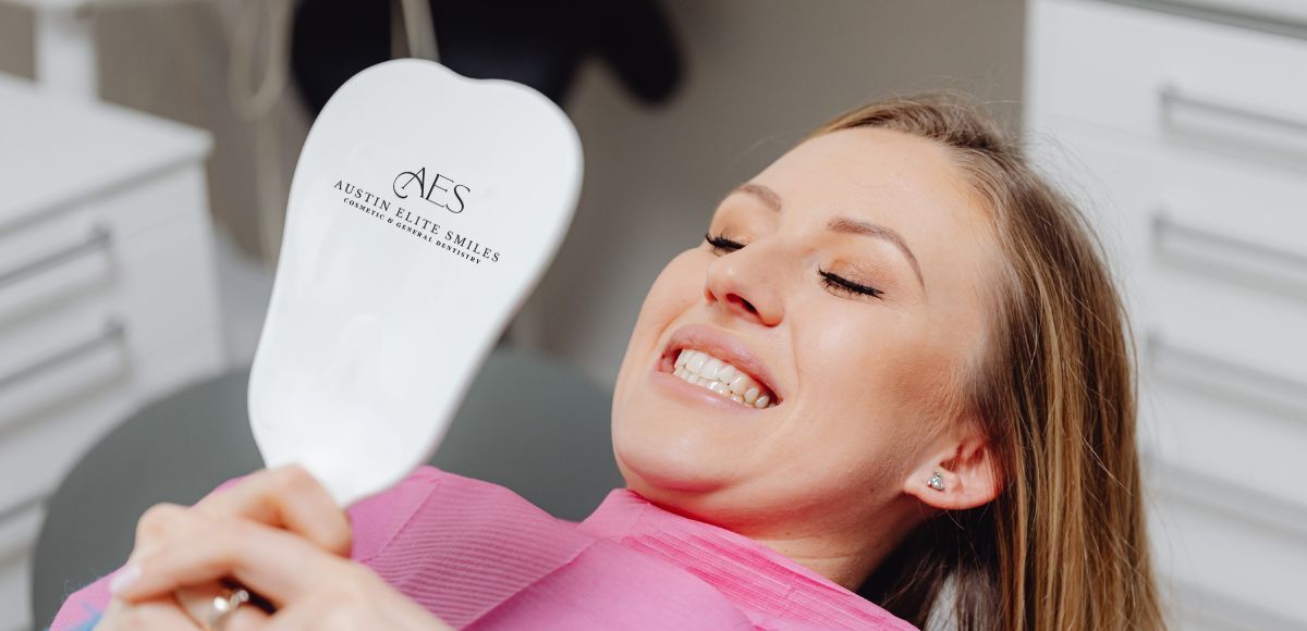 Woman smiling in a dental chair, holding a white handheld mirror in a bright clinic