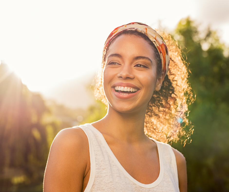 Woman with curly hair smiles, wearing a white tank top and a patterned headband, outdoors with sunlight.