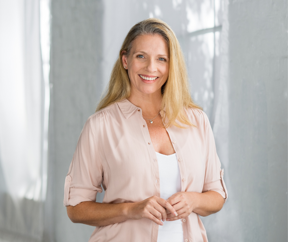 Woman with long blonde hair, smiling. Wearing a light pink button-down shirt over a white top. Standing indoors.