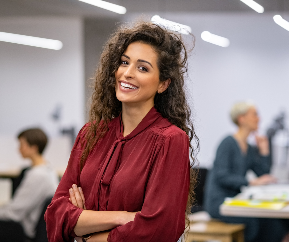 Woman with long curly hair smiles, arms crossed, wearing a maroon blouse in an office setting.