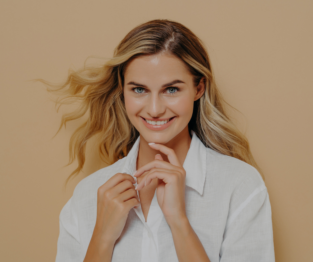 Woman with blonde hair smiles, buttoning a white shirt, beige background.