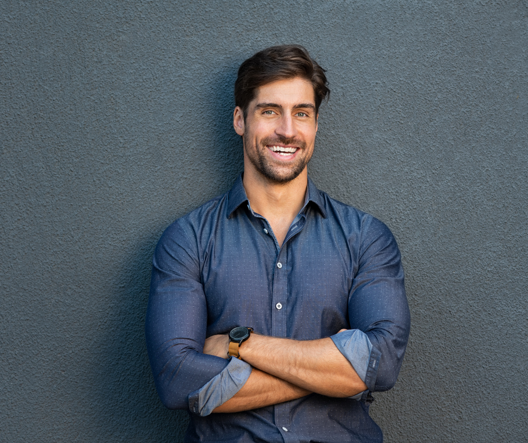 Man in blue button-down shirt smiles, arms crossed, in front of a gray wall.