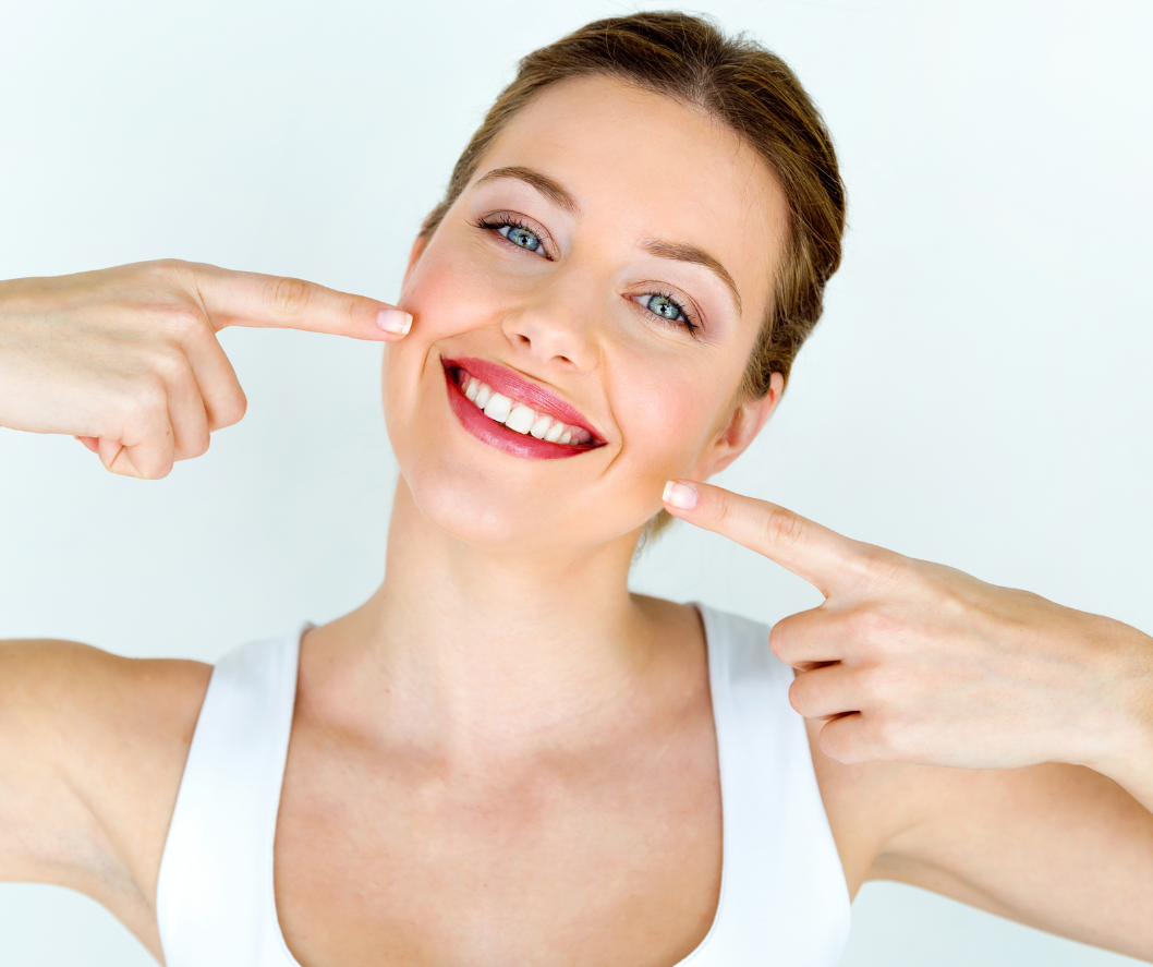 Woman with a bright smile, pointing to her cheeks, white tank top, light background.