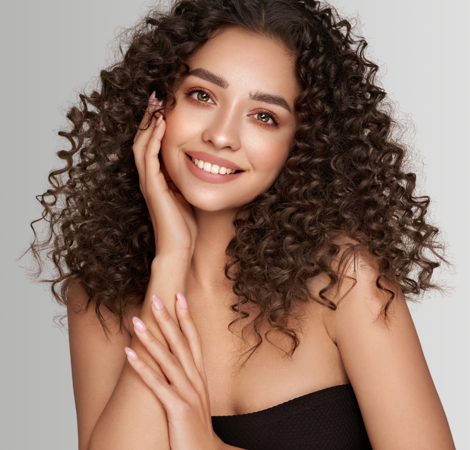 Woman with curly brown hair smiling, touching her face. She wears a black strapless top, against a gray background.