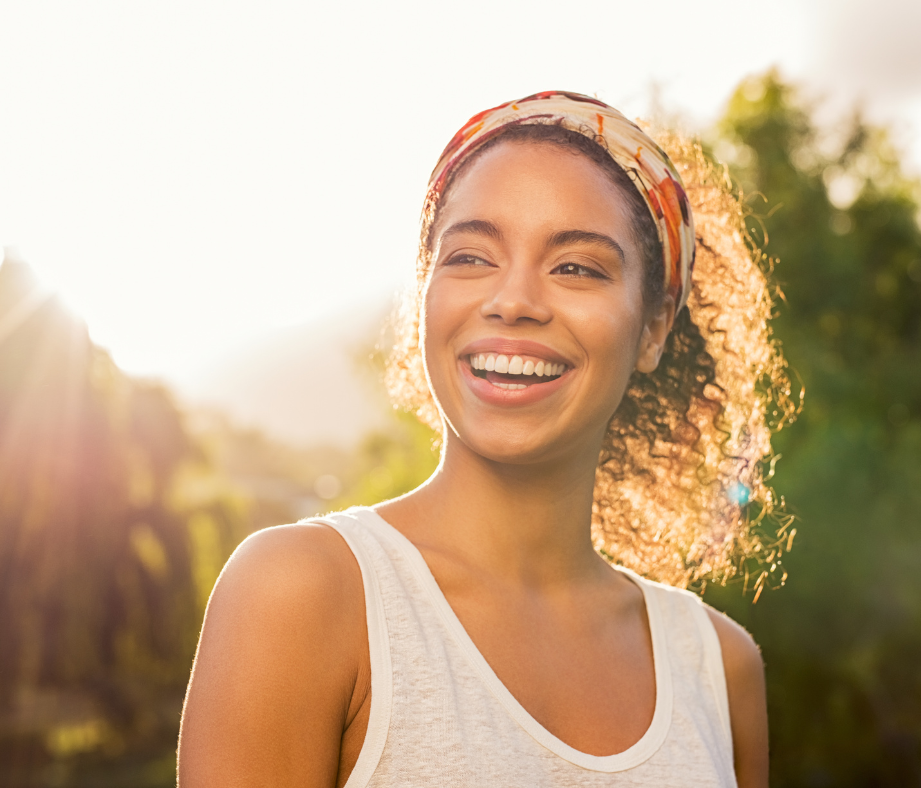 Woman smiles outdoors in sunlight, wearing a headband and a white tank top.