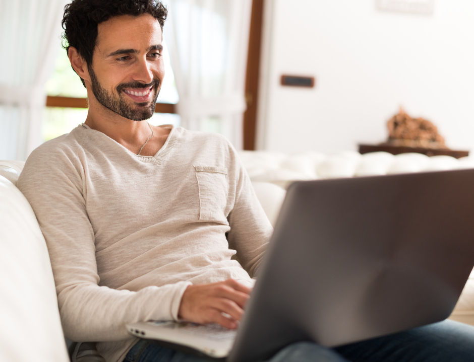 Man smiling, using a laptop while seated on a white couch. Indoor setting, natural light.