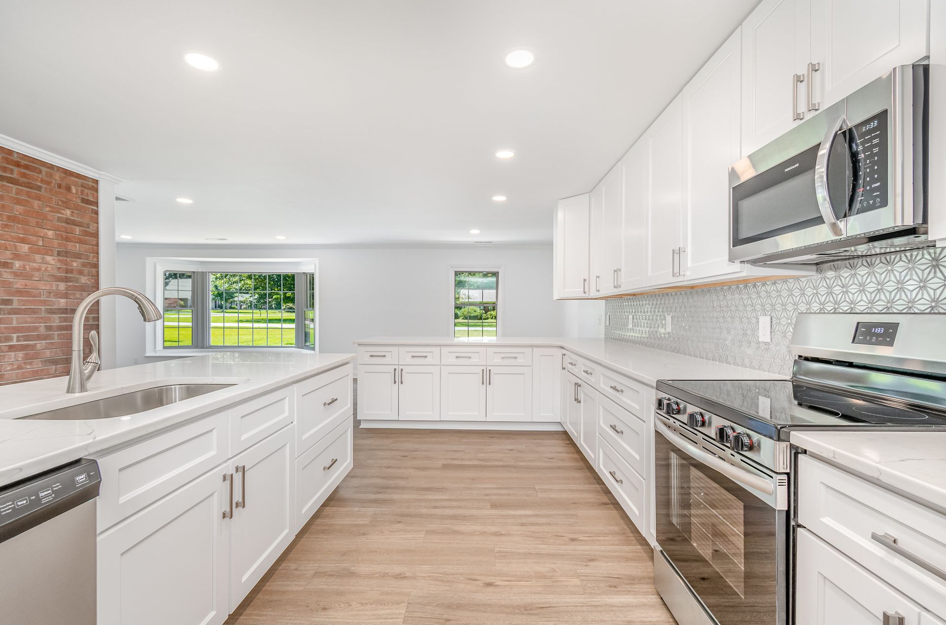 Modern white kitchen with stainless steel appliances, light wood floor, and brick wall.