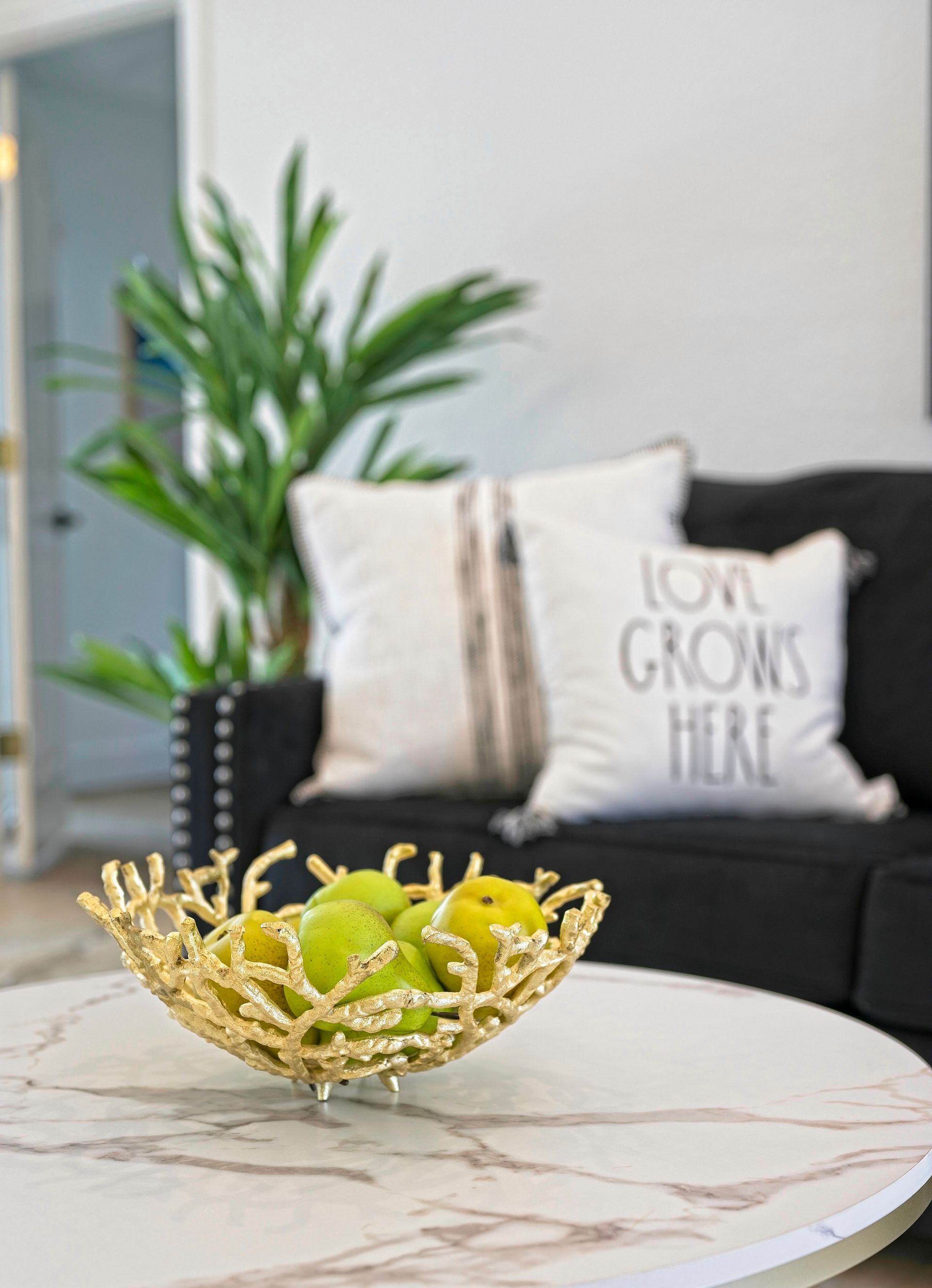 Gold decorative bowl with green apples on a marble table; black sofa with pillows in the background.