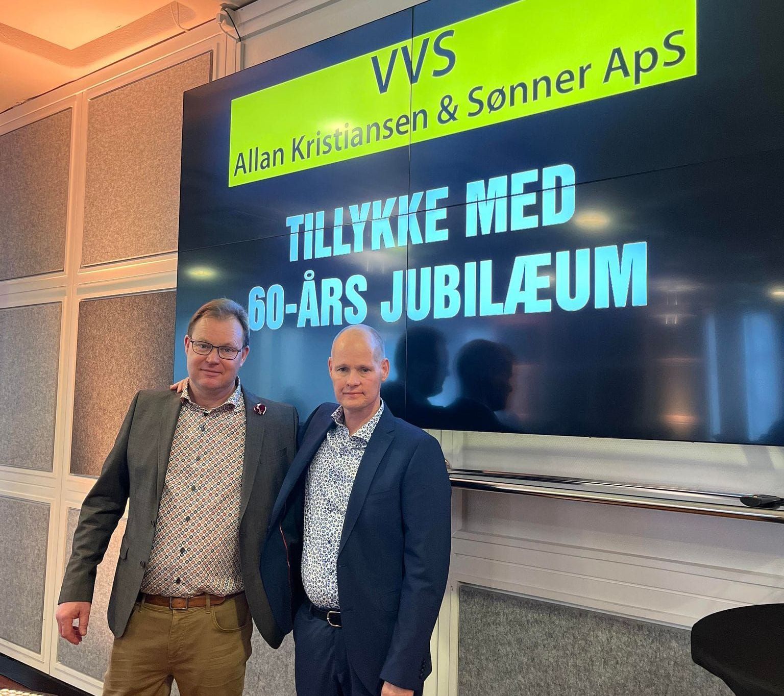 To mænd, der står foran et skilt, hvor der står 60 års jubilæum
