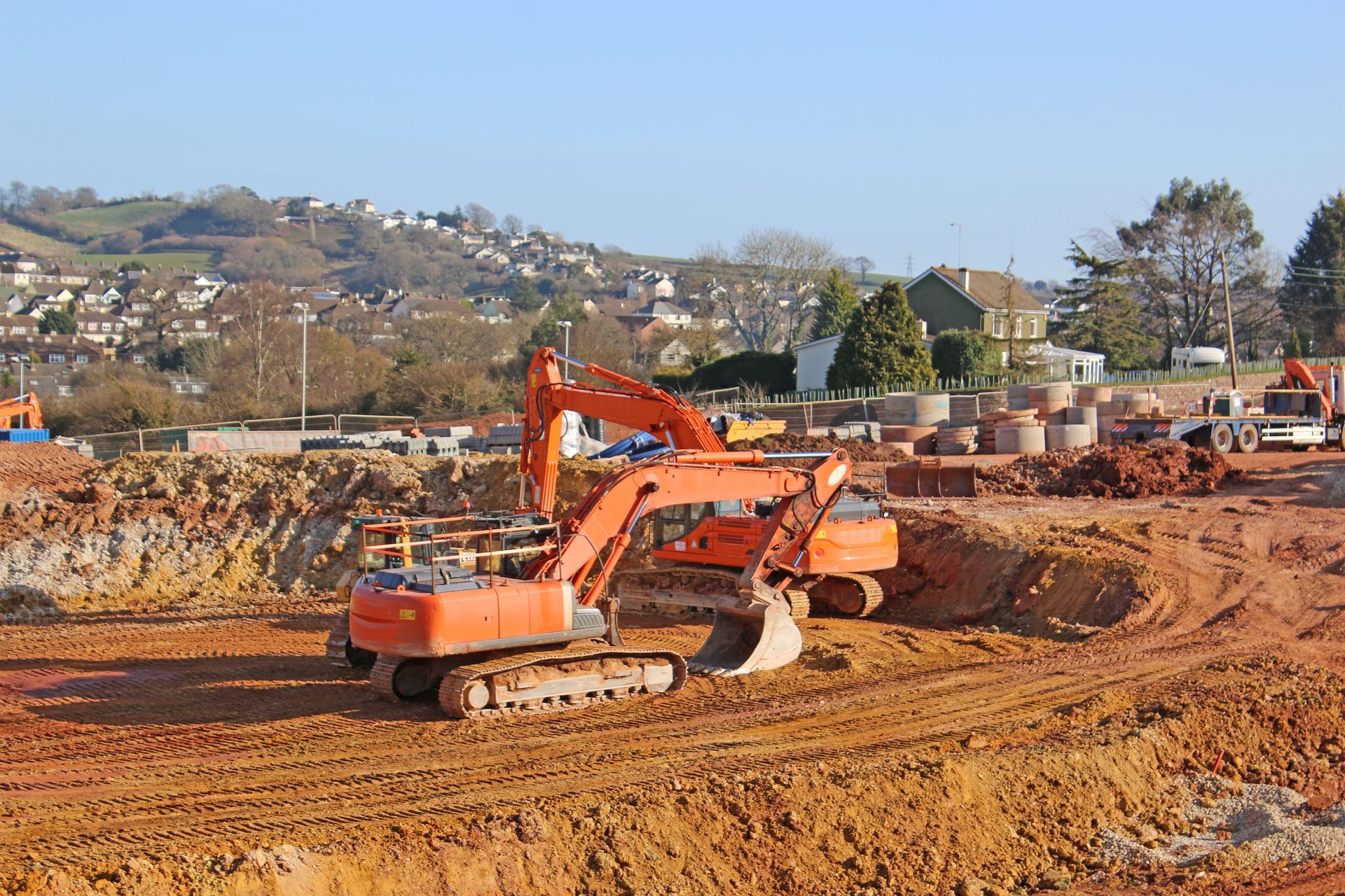 Two orange excavators on a construction site, digging into reddish-brown earth. Houses and green hills in the background.