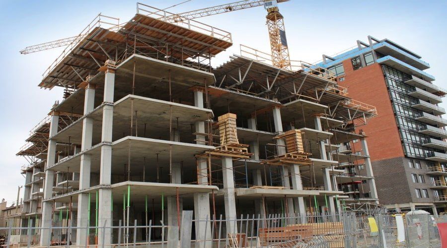 A concrete high-rise building under construction with a large crane, alongside a finished apartment building.