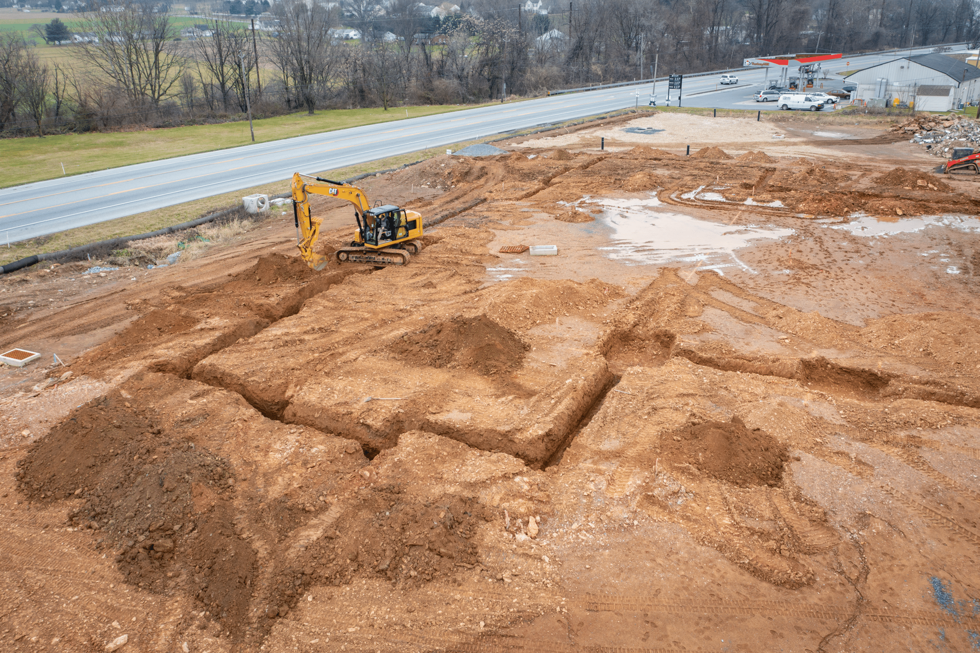 An aerial view of an excavator digging trenches for a building foundation in a construction site next to a road.