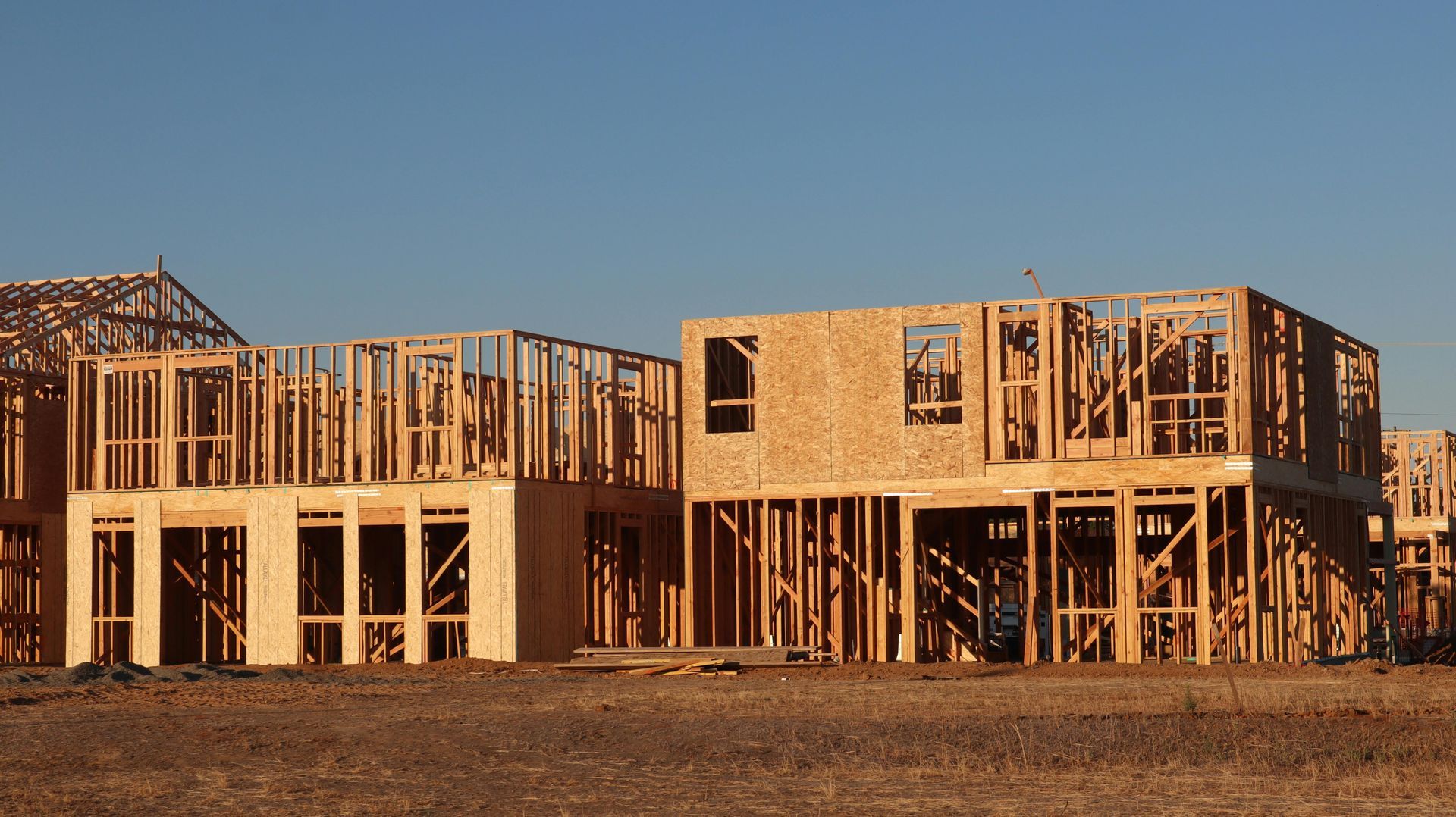Two unfinished wood-framed houses under a clear blue sky on a dusty construction site.