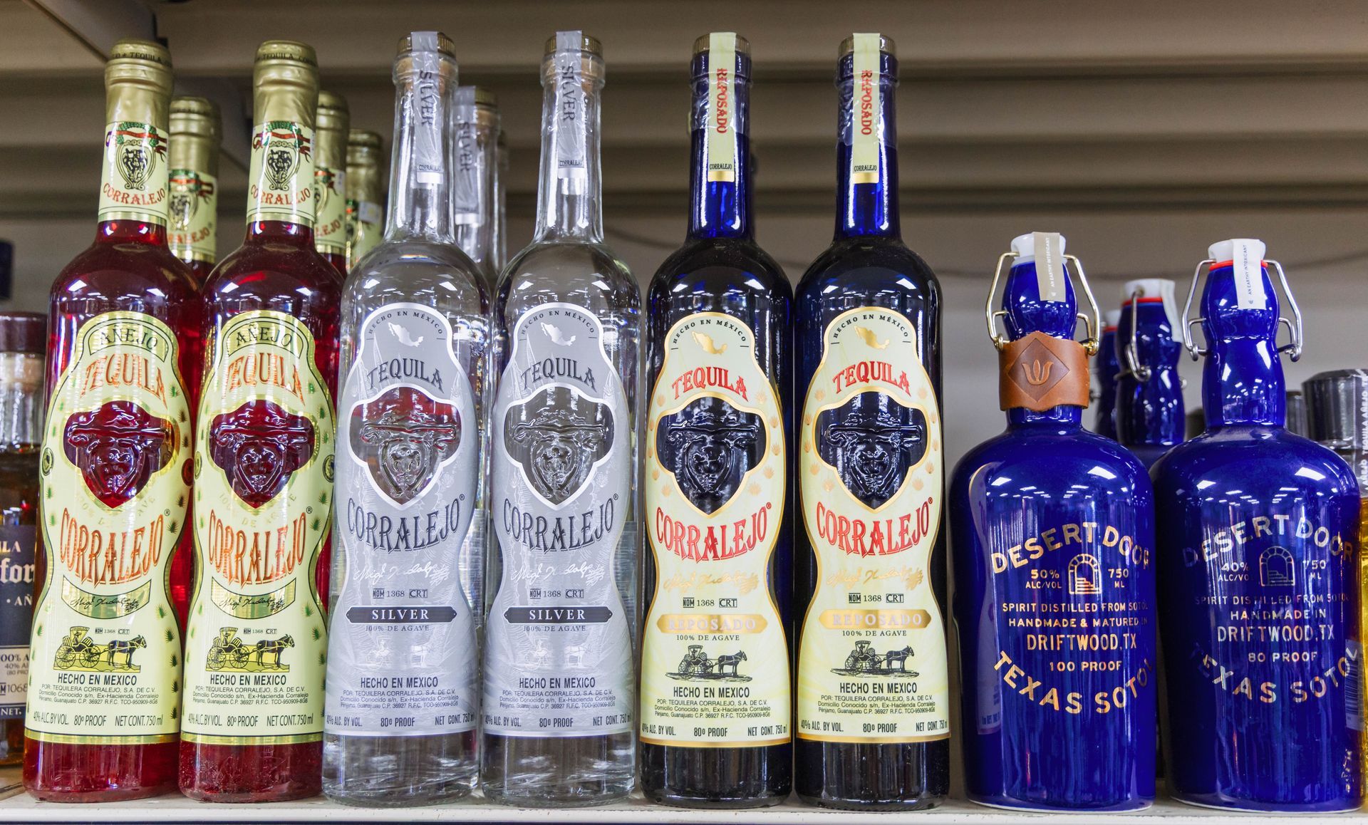 Shelf lined with tequila bottles; various colors and labels, some with swing-top closures.