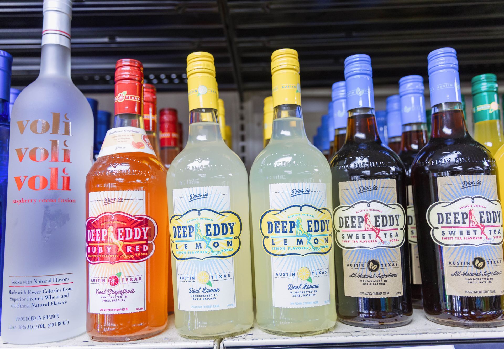 Bottles of alcoholic beverages on a shelf.  Brands include Voli vodka and Deep Eddy flavored vodkas like lemon and cranberry.