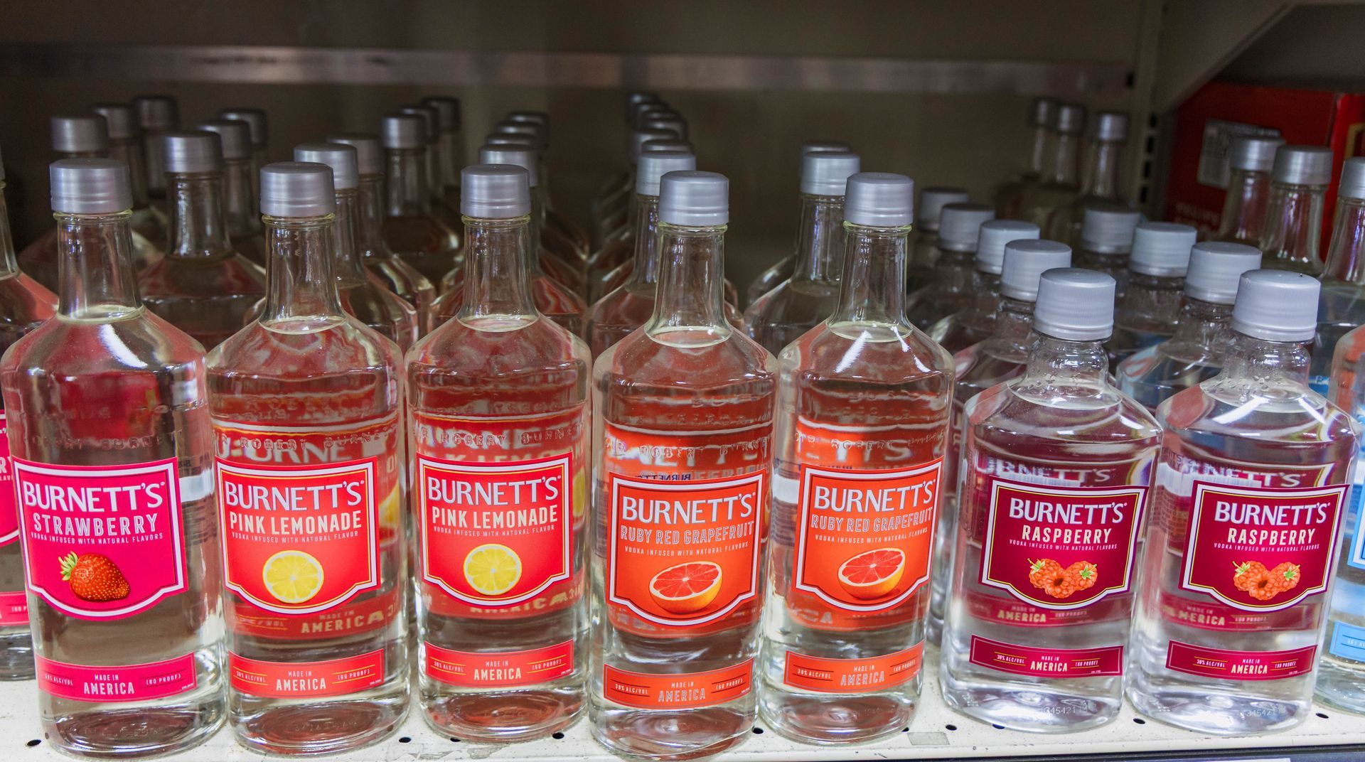 Bottles of Burnett's flavored vodka on a shelf, including raspberry, orange, and peach, with gray caps.