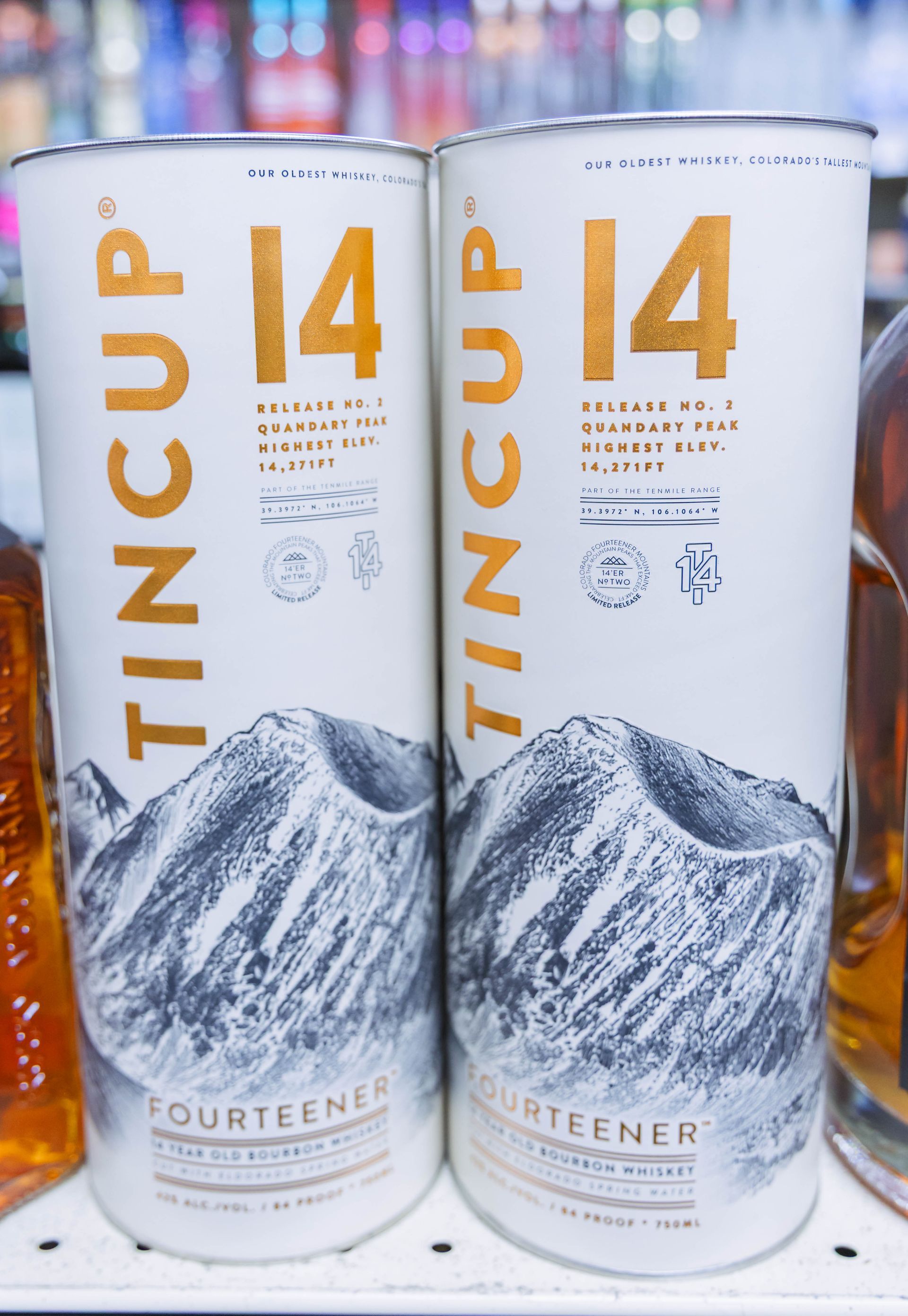 Two cylindrical white TINCUP I4 whiskey bottles with gold text and mountain illustration.
