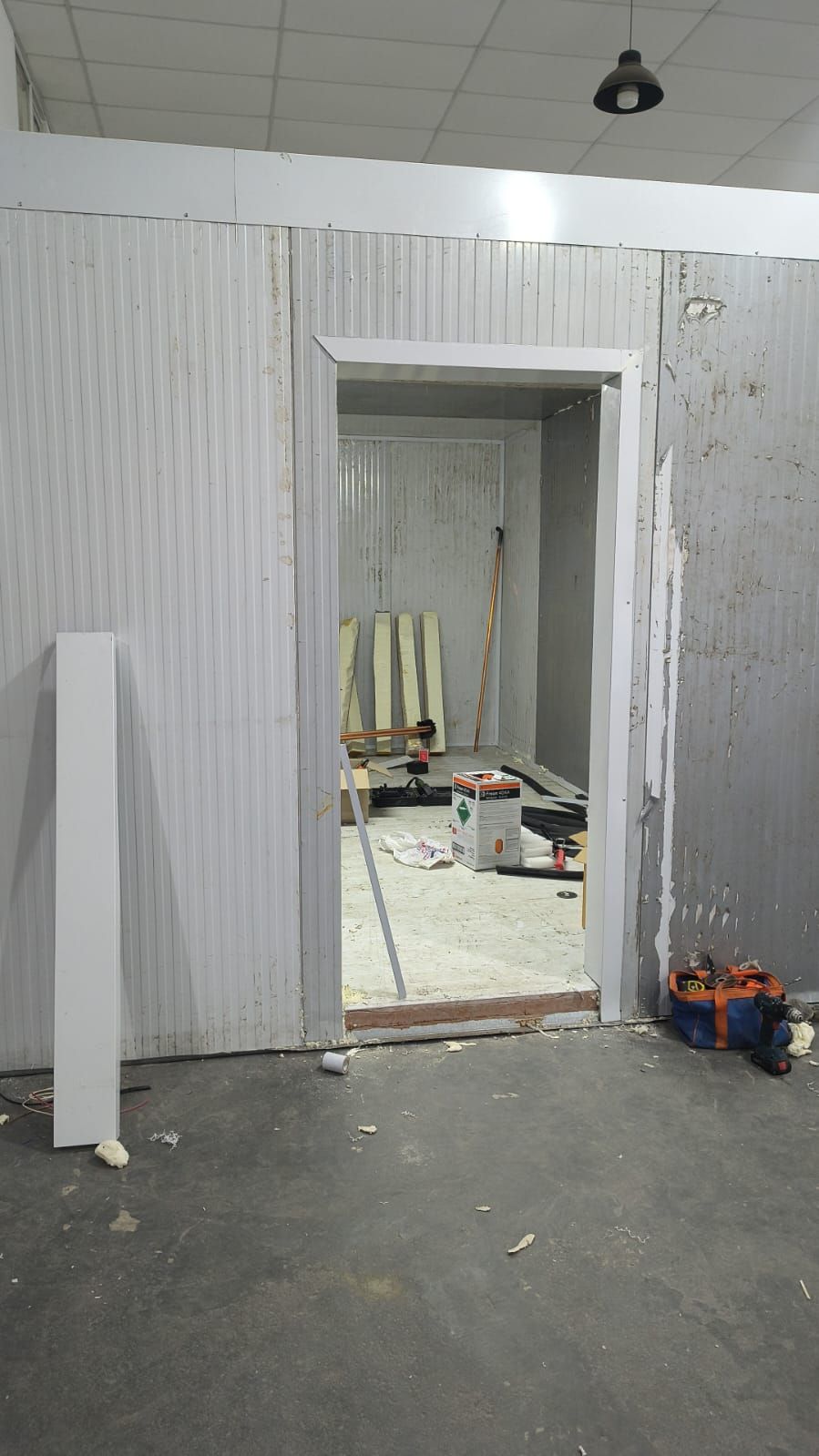 Habitación parcialmente reformada con paredes de chapa ondulada, puerta abierta y herramientas de construcción en el suelo.