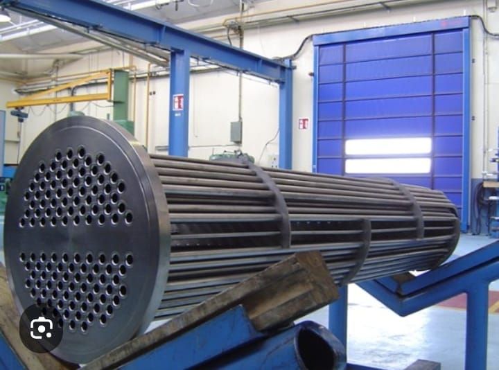 Gran haz de tubos metálicos dentro de un taller industrial, cerca de maquinaria azul y una puerta azul cerrada.