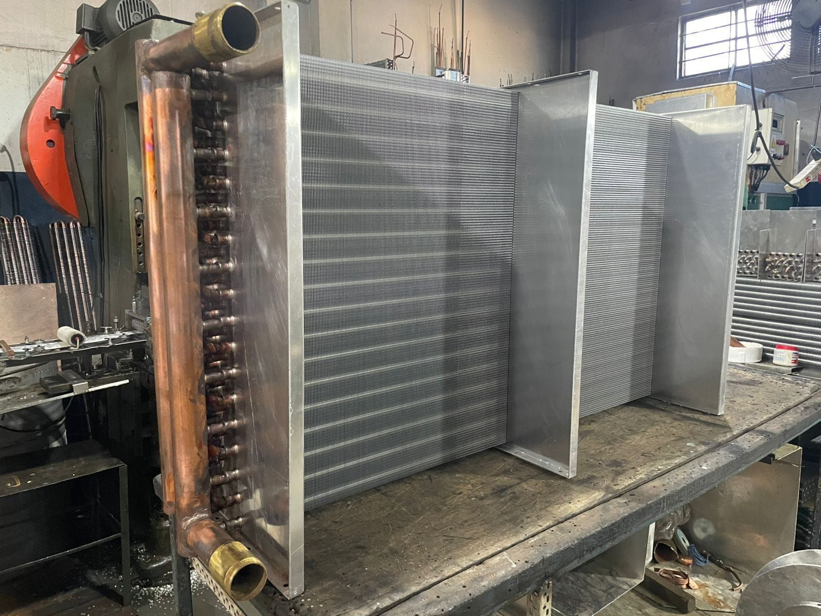 Intercambiador de calor industrial de metal sobre un banco de trabajo, con tuberías de cobre y aletas grises en un taller.