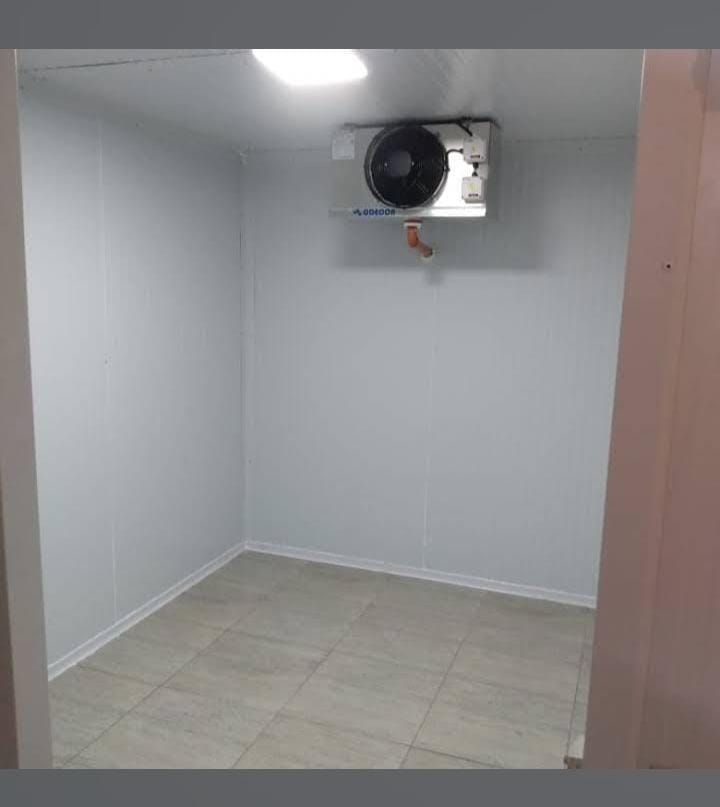Cámara frigorífica vacía con paredes grises, suelo de baldosas y un ventilador de pared cerca del techo.