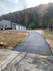 Newly paved asphalt driveway leading to a grey building with garage doors, set in a rural area.