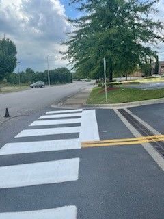 Crosswalk with white stripes, yellow lines, and a tree on a corner of a road under a cloudy sky.