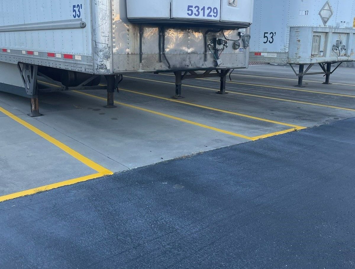 Semi-truck trailers in a parking area with yellow lines; asphalt and concrete surfaces.