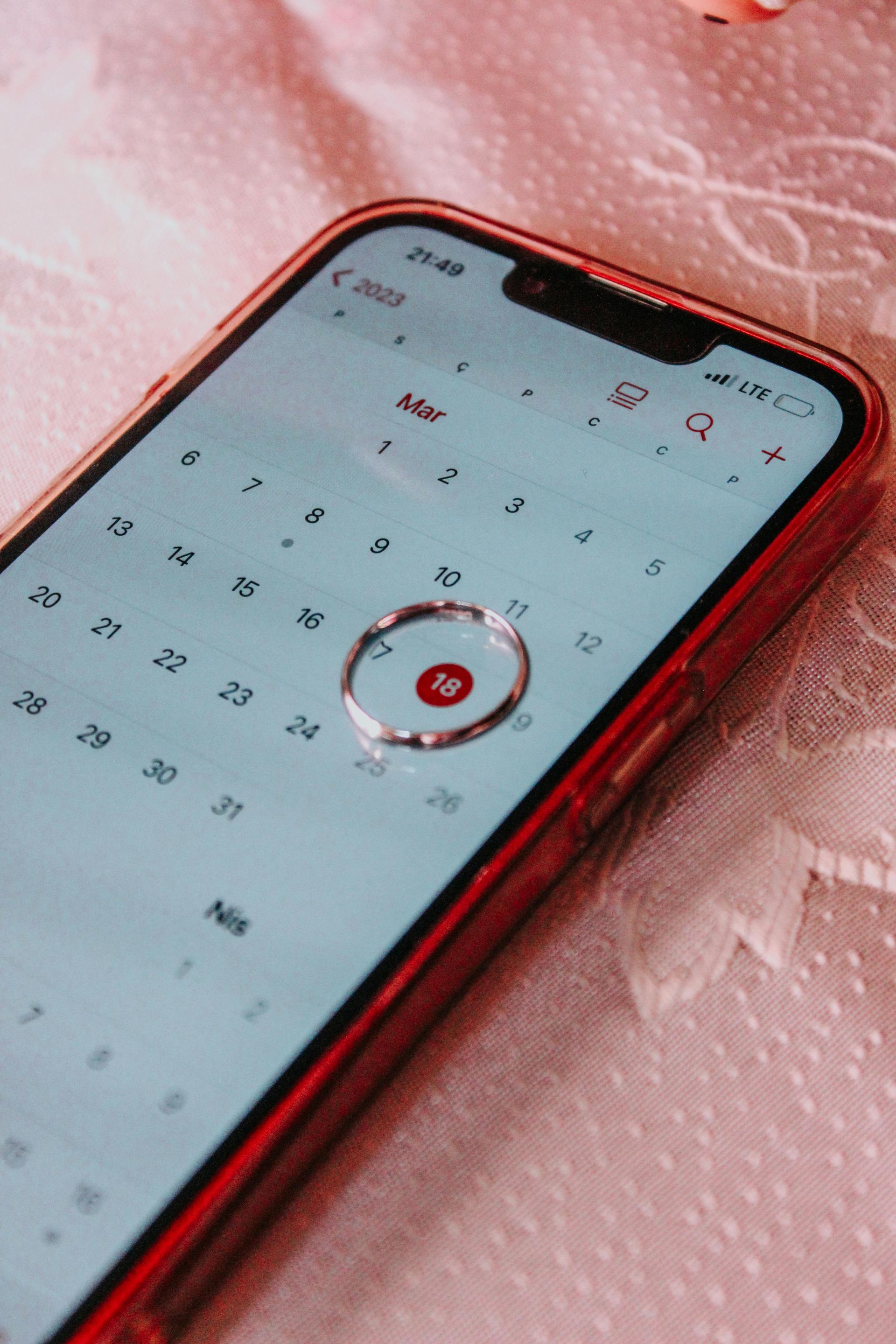 A smartphone displays a calendar app with the 18th circled, resting on a textured pink surface.