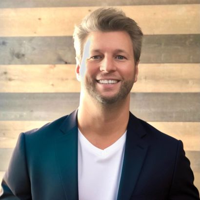 A man in a suit and white shirt is smiling in front of a wooden wall